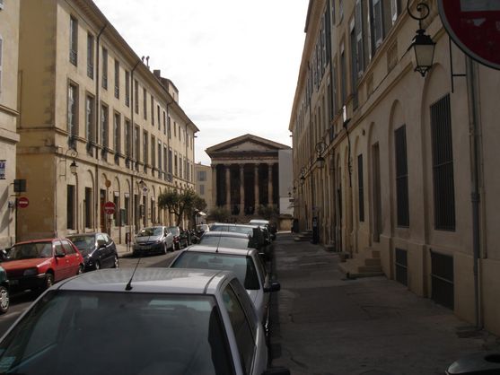 A narrow street full of parked cars. Unexpectedly, there is a Roman temple at the end of the street.
