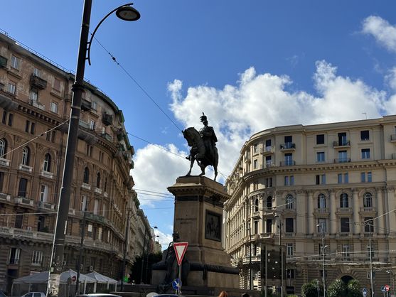 A tall stone pedestal in the middle of a roundabout. On top is a bronze statue: a figure on horseback in uniform with a massive plumed helmet. Text on the pedestal says A VITTORIO EMANUELE II MDCCCXCVI. Behind the statue are some grimy but glamorous buildings, just a few storeys high.