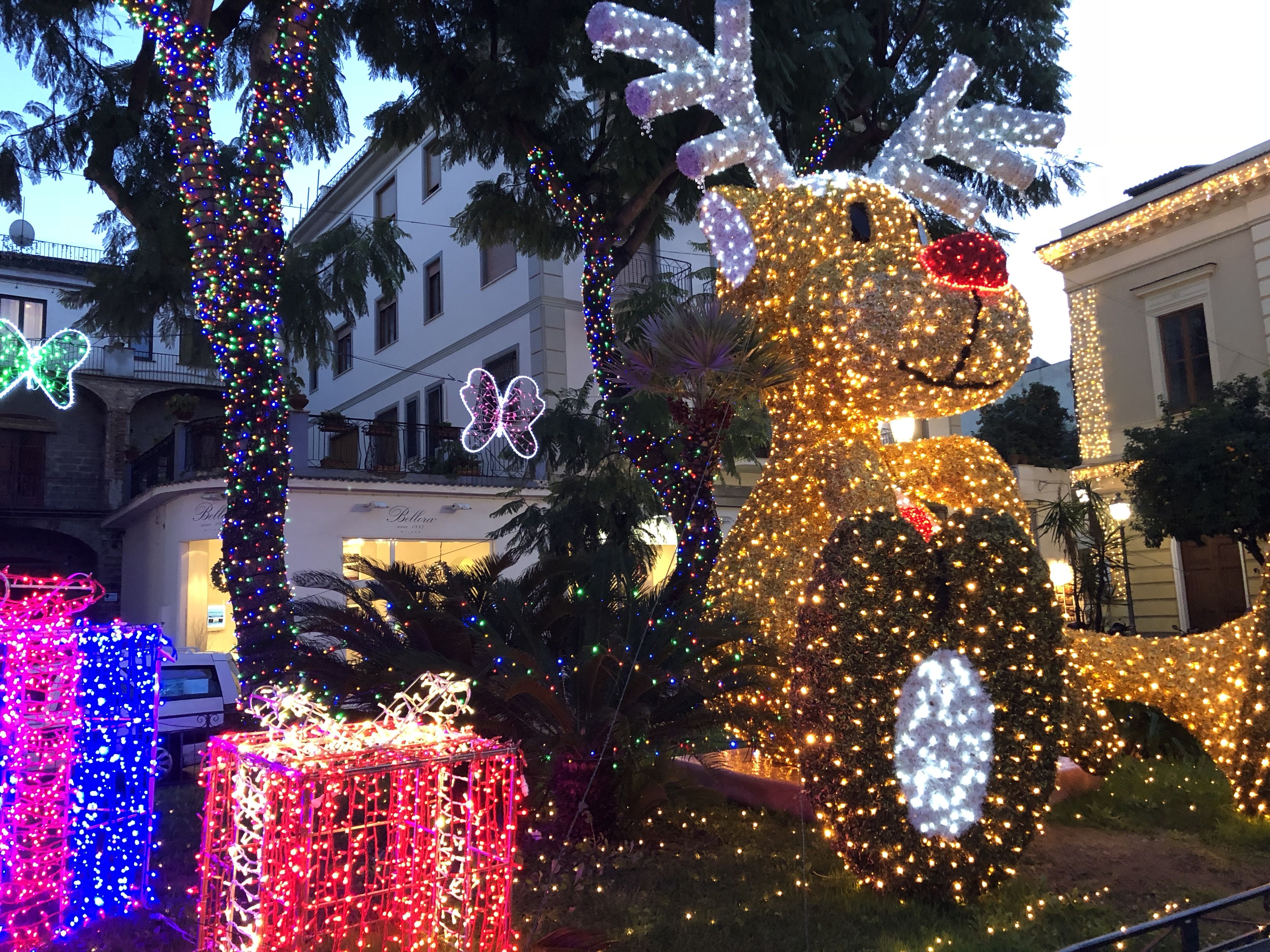 A square in the middle of Sorrento. Trees are festooned  with Christmas lights, and next to theme is a giant cartoon reindeer made of wire and Christmas lights, next to giant Christmas presents