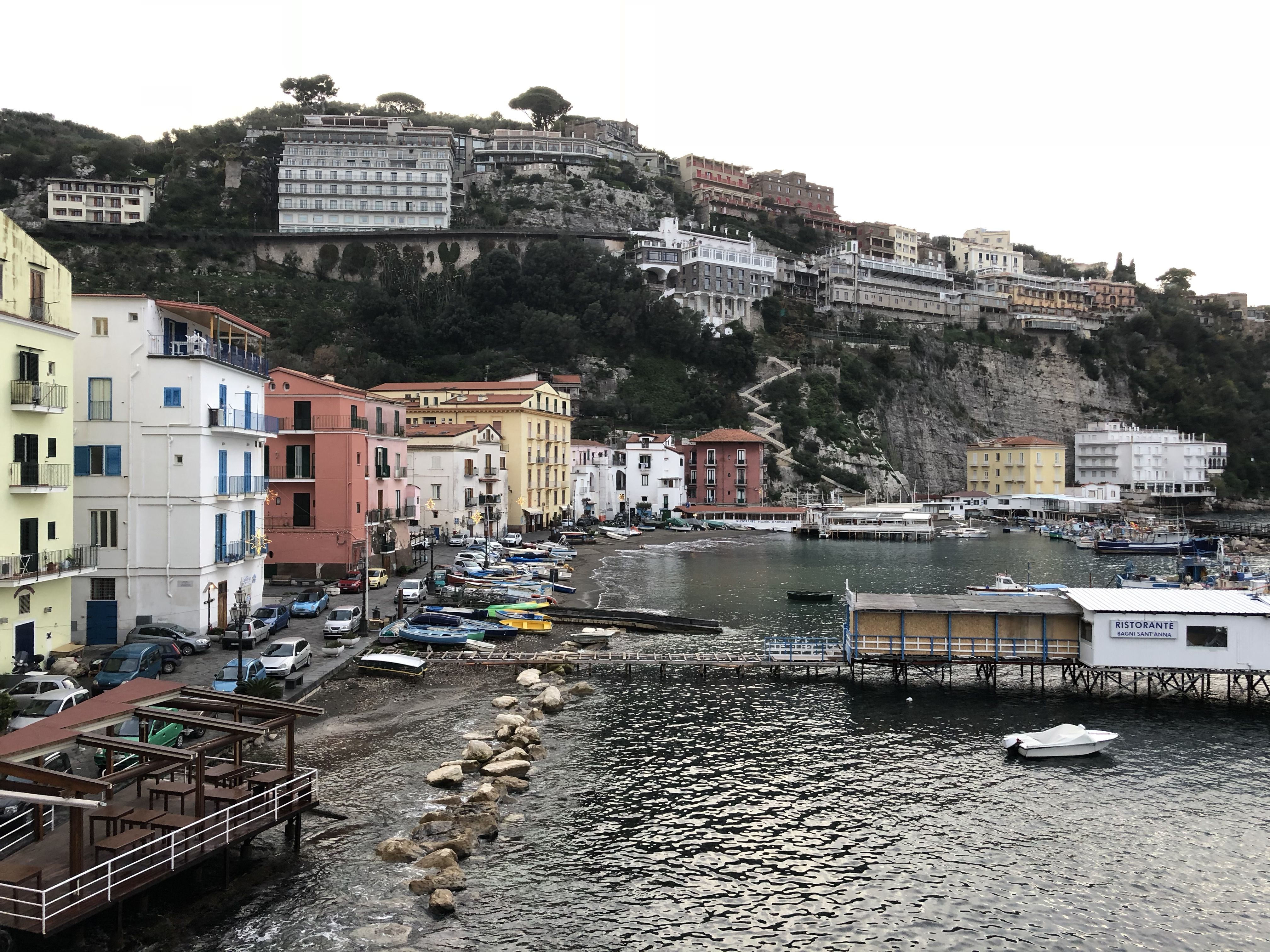 Down at the waterfront. There's a pier with a little white wooden  restaurant at the end. Boats in the water. Just back from the water, a row of hotels. And looming above, at the top of a cliff, a row of hotels and apartment buildings.