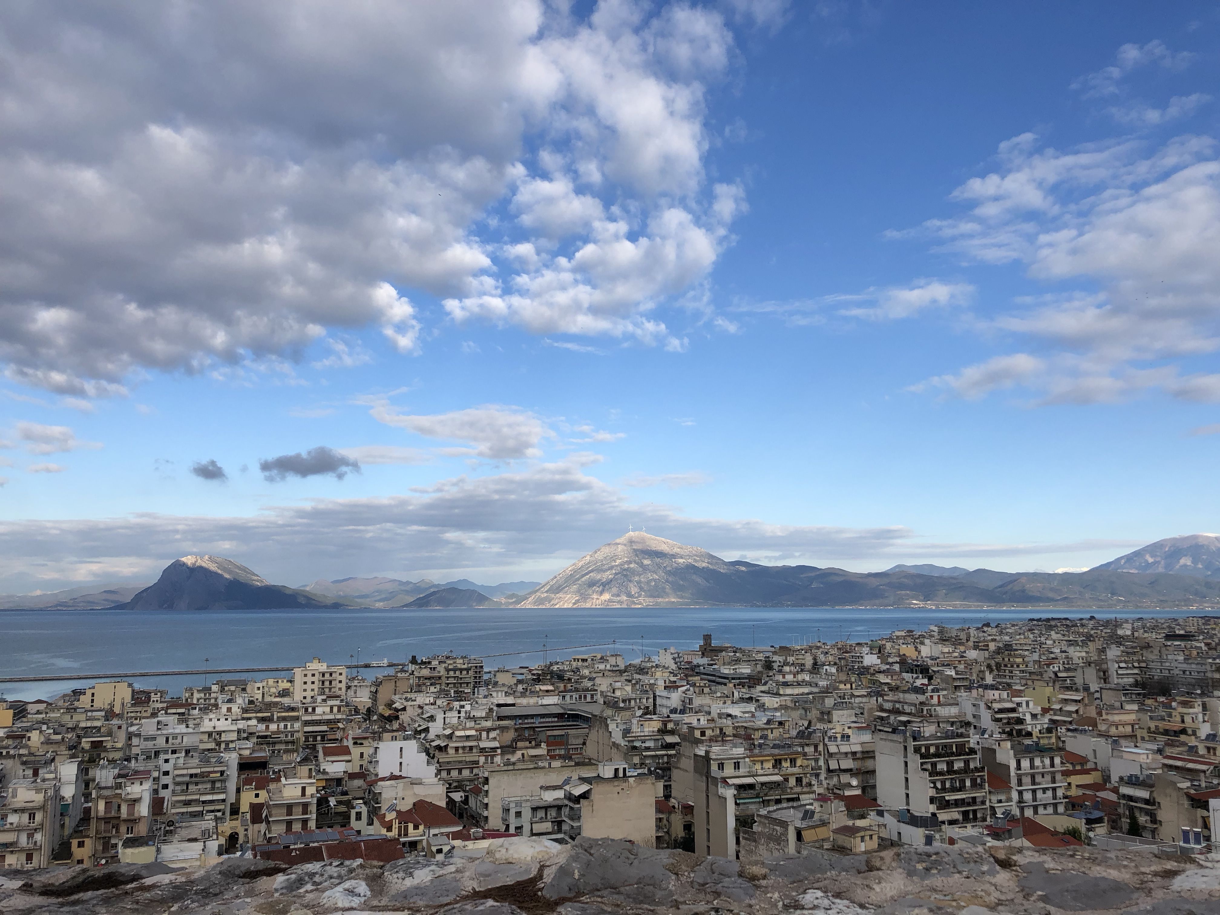 Looking out over Patras, in shadow. Across the water are lightly wooded mountains.