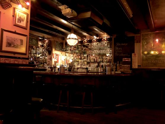 An old-fashioned wood-panelled bar with shelves of bottles in the background, lit by a dim yellow light.