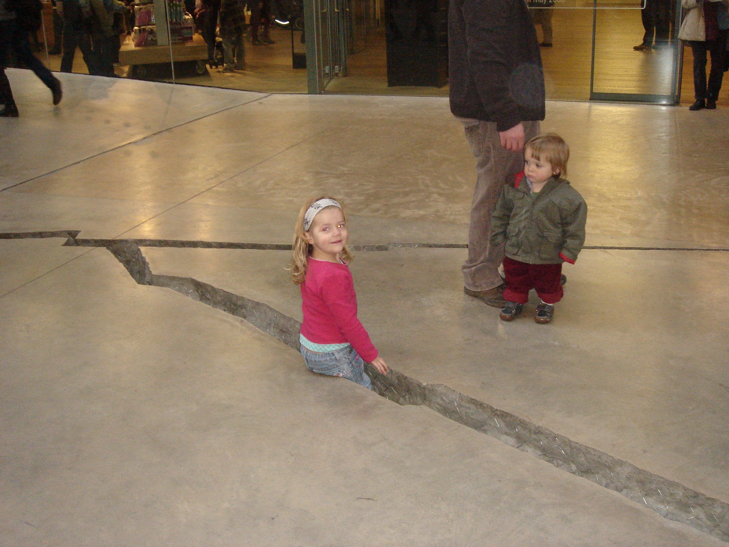 A small girl is standing in a crack in a concrete floor, looking at us. A tiny toddler is holding a parent's hand and looking over at her.