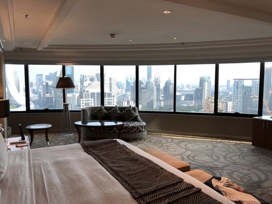 An expensive hotel bedroom. A corner of the bed is visible, but the most striking thing is the view of a cluster of skyscrapers through the panoramic windows. There's a couch in front of the windows, facing absolutely the wrong way.