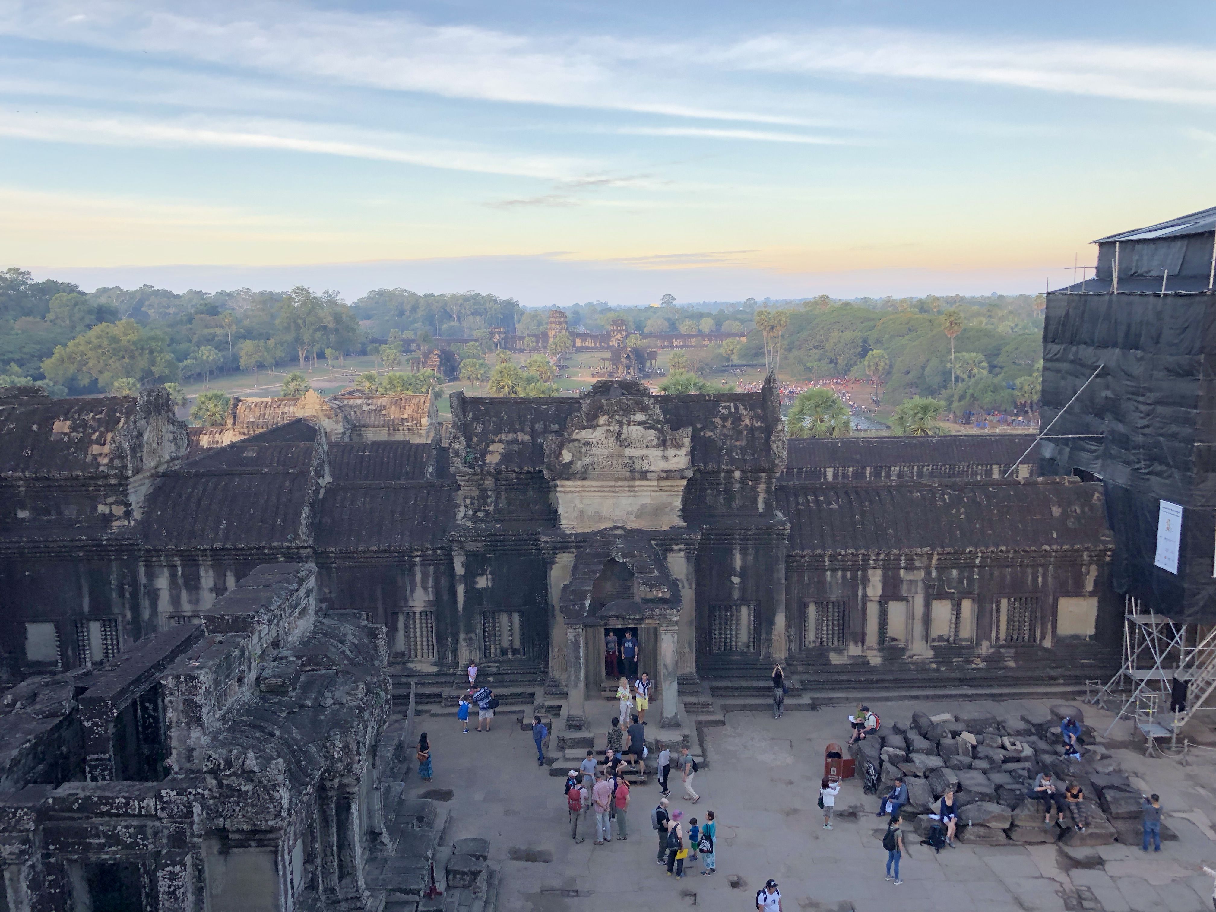 Looking out at a courtyard in Angkor Wat. The wall facing us has a  door flanked with pillars. There are tourists milling round. More sandstone structures are visible in the distance.