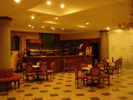 A nineties-style hotel lobby, with a bar and lots of table settings in front of it. The floor is tiled and the light is very yellow.