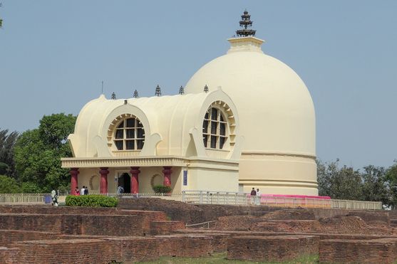 A small temple with a roof shaped like half a cylinder and a round window on the wall facing us. A big hemispherical stupa stands behind it. They are both painted dark cream.