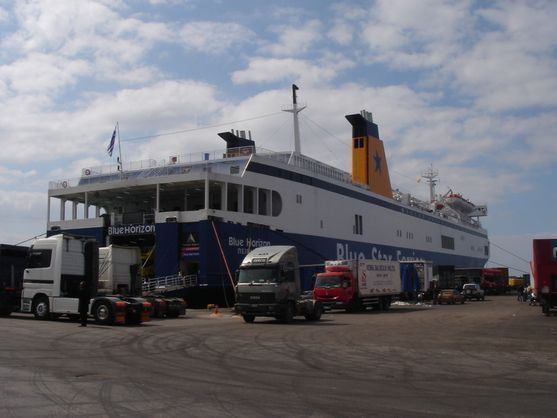 A large white and blue Mediterranean ferry marked Blue Star Ferries is in dock, with some large trucks in front of it.