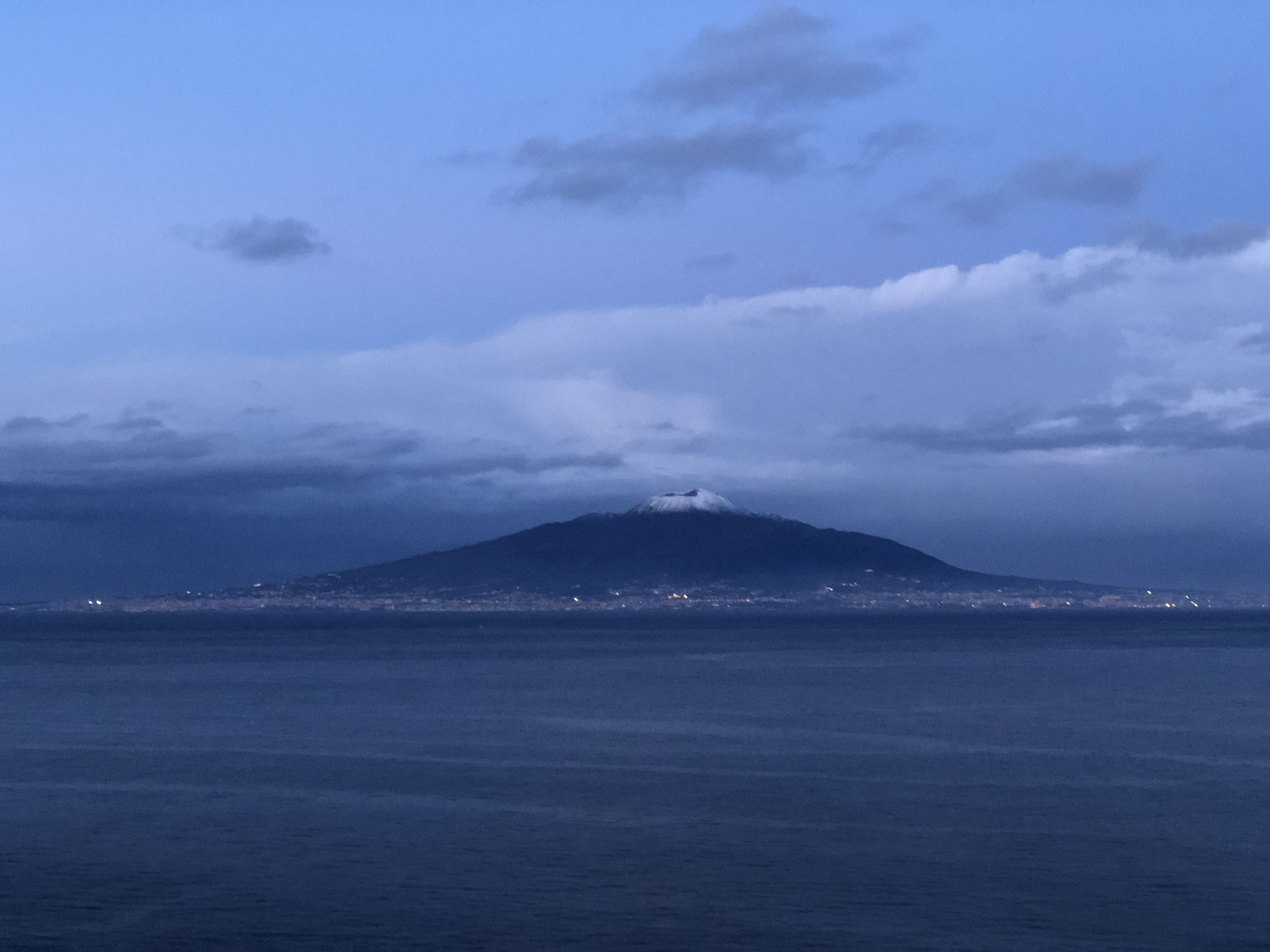 It's dusk, and across the Bay of Naples stands Mount Vesuvius. At the foot of the mountain a few scattered lights can be seen.