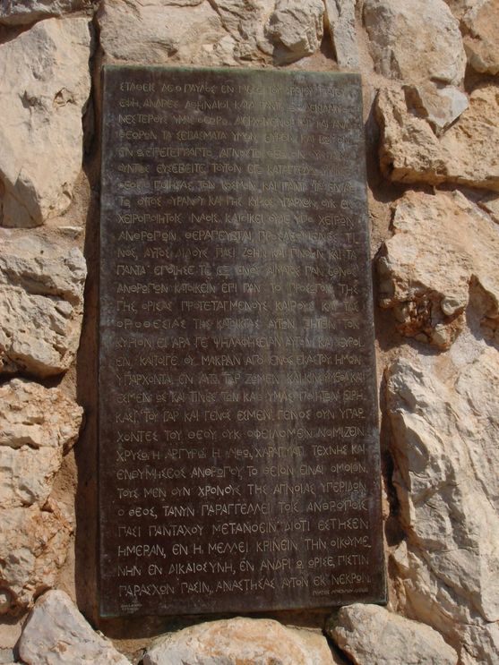 Closeup of a bronze plaque set into a stone surface. The plaque is inscribed with Greek capital letters.