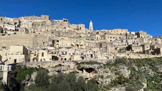 The Sassi: rows and rows of yellow stone buildings cover the side of   a hill. There's a road below them, and below the road the scrubby hillside can be seen. It's a beautiful sunny day.
