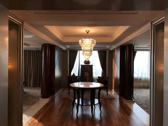 A tastefully dark entry hall, with pillars in the corners, a circular table in the middle, and a heavily-curtained window at the end. There is a massive chandelier hanging from the ceiling.