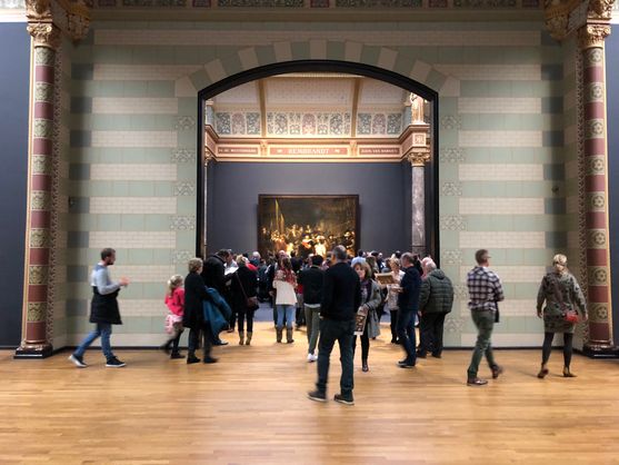 We're inside the Rijksmuseum in Amsterdam. Visitors are walking around in all directions. Behind them is a large arch in the wall with the name Rembrandt inscribed above it; behind that we can just make out The  Night Watch.