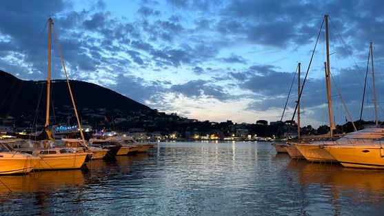 Looking out across a small harbour in the evening.  There are some small yachts in the foreground and a wooded hill  in the background. The sky is dark with blue clouds,  and the sun has just set.