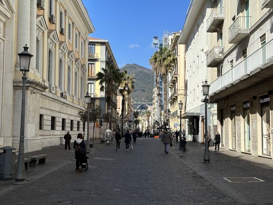 Looking along a cobbled street in an Italian town. There are a few pedestrians walking along the street. On either side of the street are the usual Italian buildings; some of them have palm trees in front of them. At the end of the street, in the distance is a wooded hill,  with a motorway running across it, supported by arches.