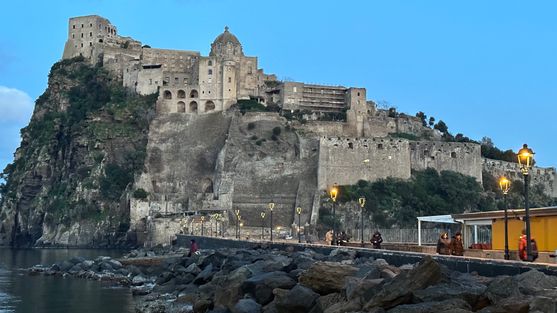 A hill juts out of the sea. There are walls built up the side facing us, and on top of those walls and the hill is a castle the same colour as the rock of the hill. A stone bridge leads from the bottom of the hill towards us.