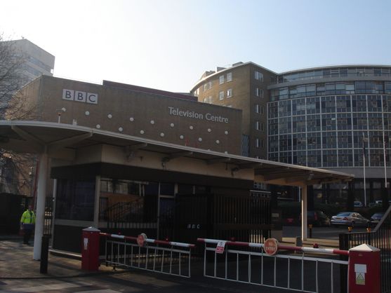 The familiar sight of the BBC Television Centre. Its a brick building with a round wall full of windows. There are traffic fences and boom gates visible in the foreground.