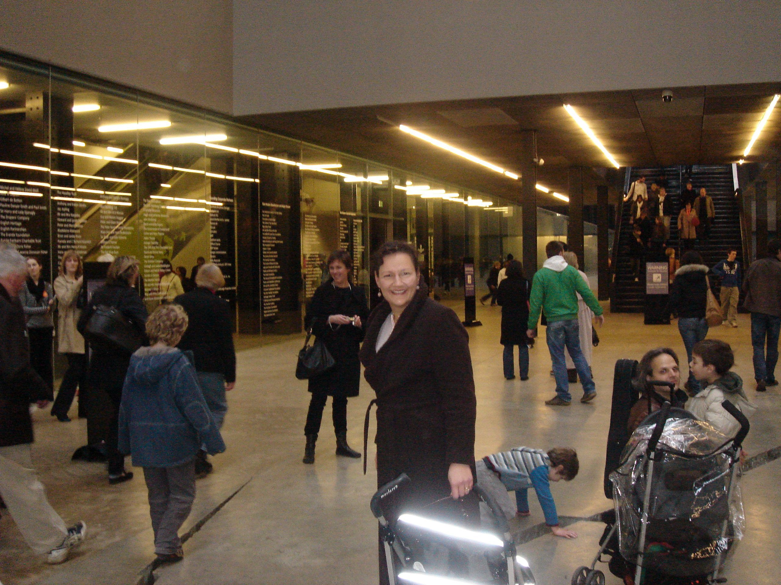 My friend Angela is standing in the foyer of the Tate Modern; she is holding onto a stroller. The foyer full of people going in different directions.