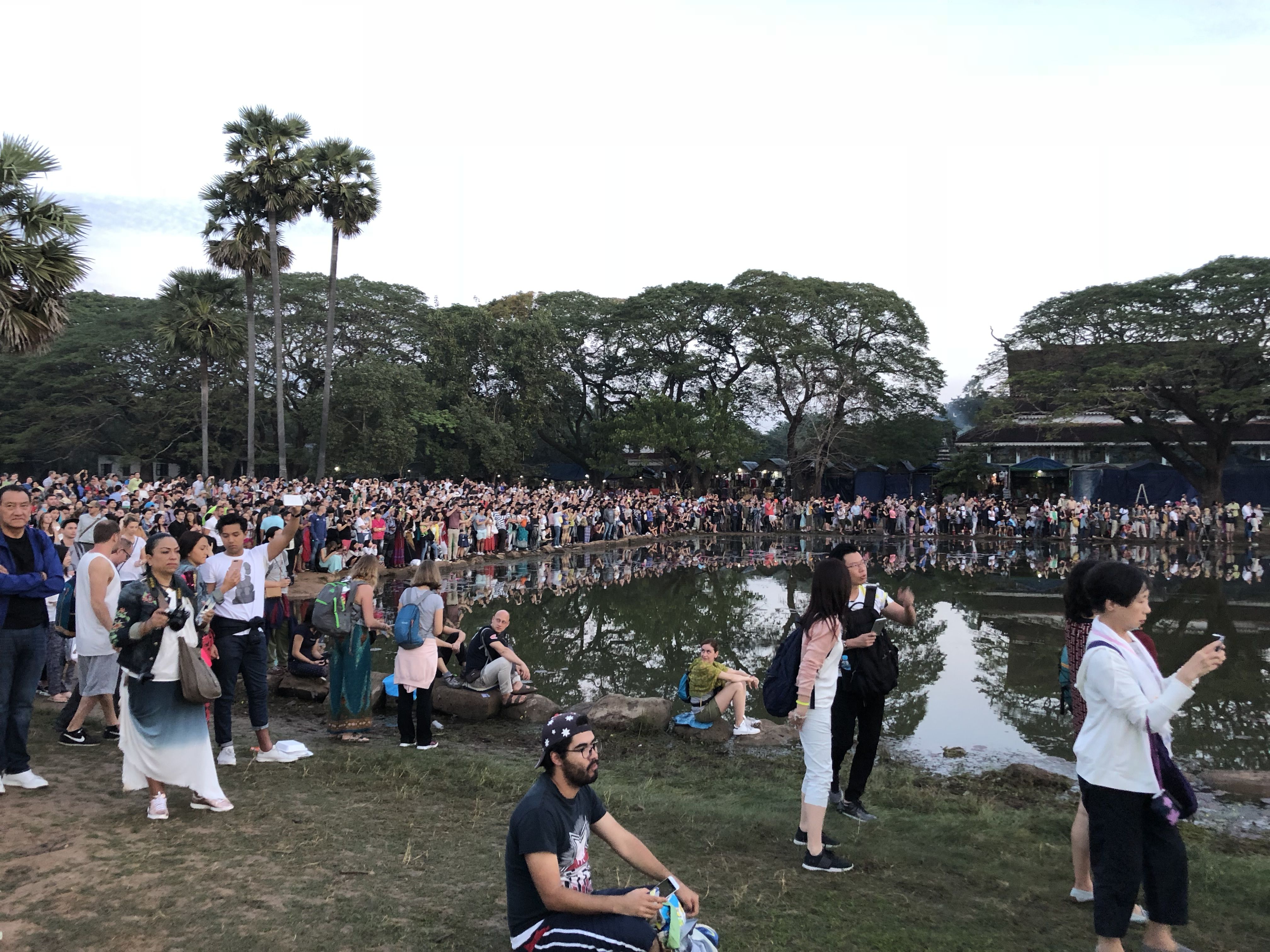 We're on the edge of the crowd which is standing around a lake.  In the distance the crowd is packed. There are trees behind them.
