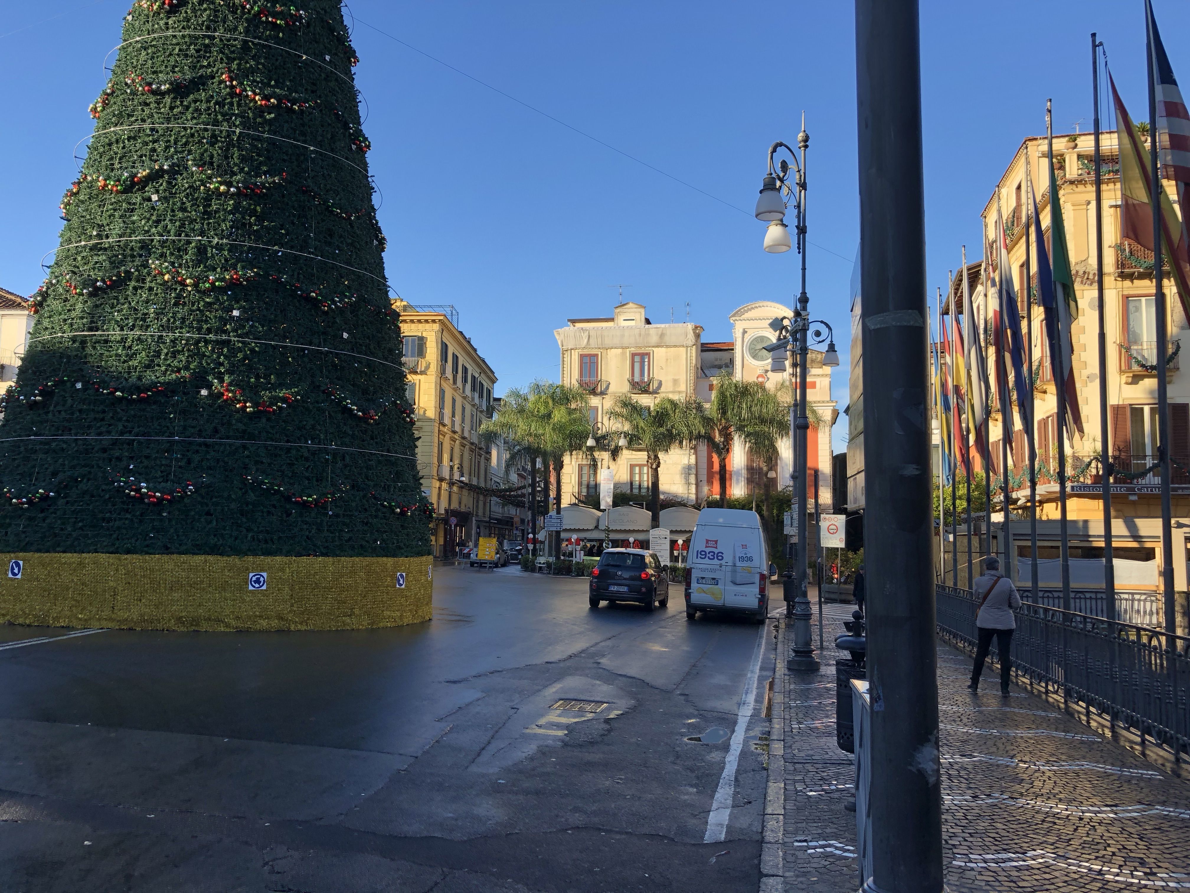 Tasso Square in the centre of Sorrento. On the left, a big conical fake Christmas tree. On the right, an old building with a row flagpoles out the front. In the centre a restaurant behind a row of palm trees.
