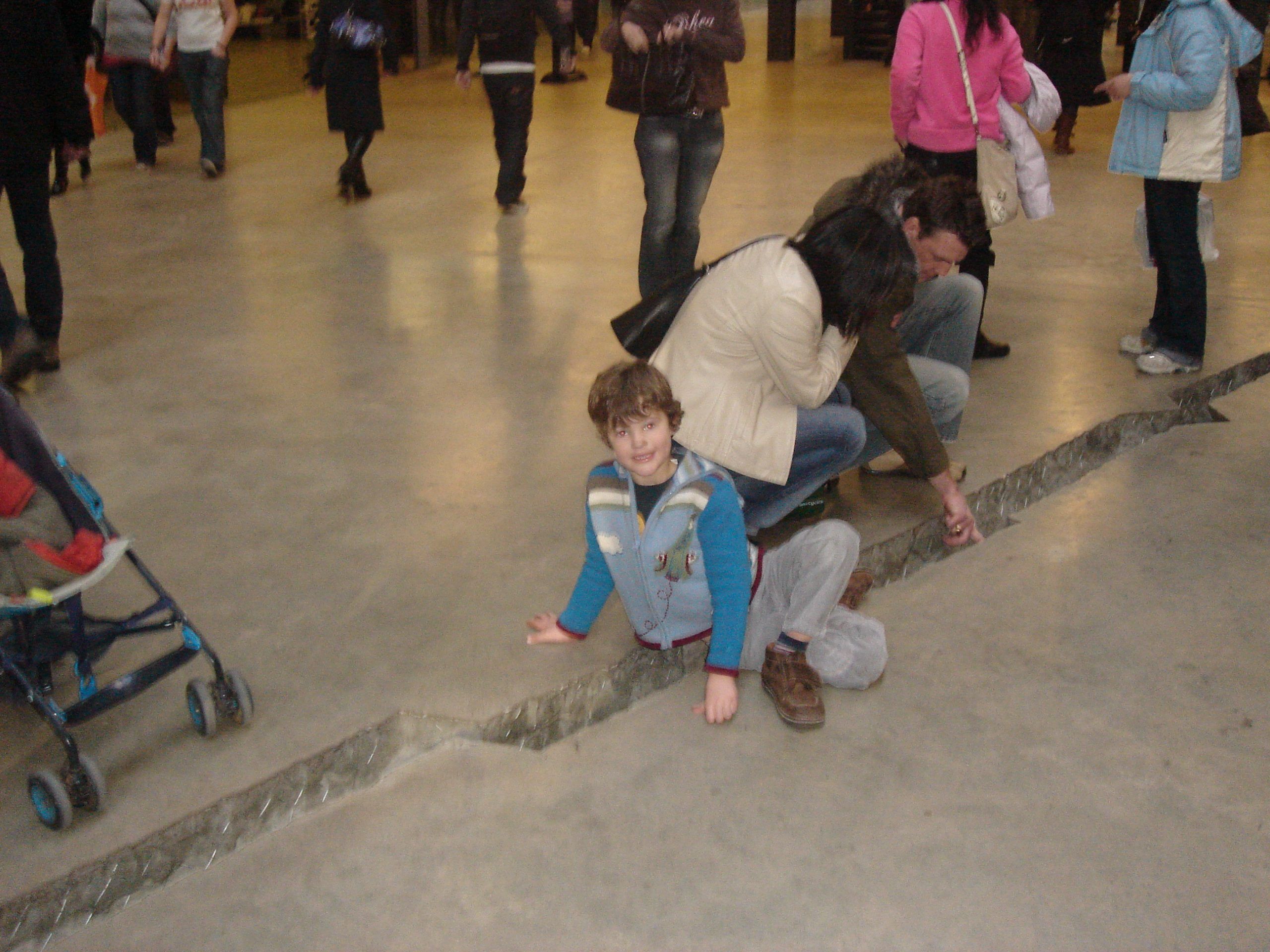 There's a crack in the concrete floor. A small boy straddles it and some adults are touching it. There are other visitors walking around.