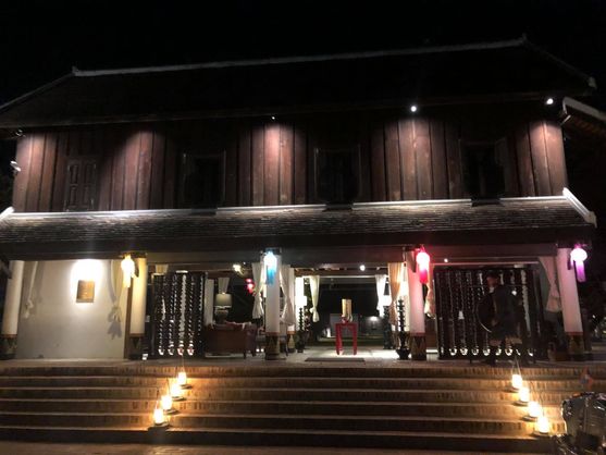 The front of a hotel. Wide stairs lead up to a pillared verandah. Each pillar is festooned with a coloured lamp. Above the awning, the walls are made of dark wood.