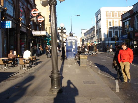 We're looking down a busy road on a sunny day.  On the left is a London Underground station; there are people sitting at tables and people walking down the footpath. In the middle of the footpath is a battered old blue Police Box.