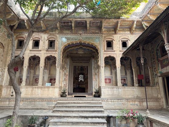 This is a sunny courtyard with a tree growing in it. The wall facing us contains ornate carvings, and a step leads up to an arched doorway.