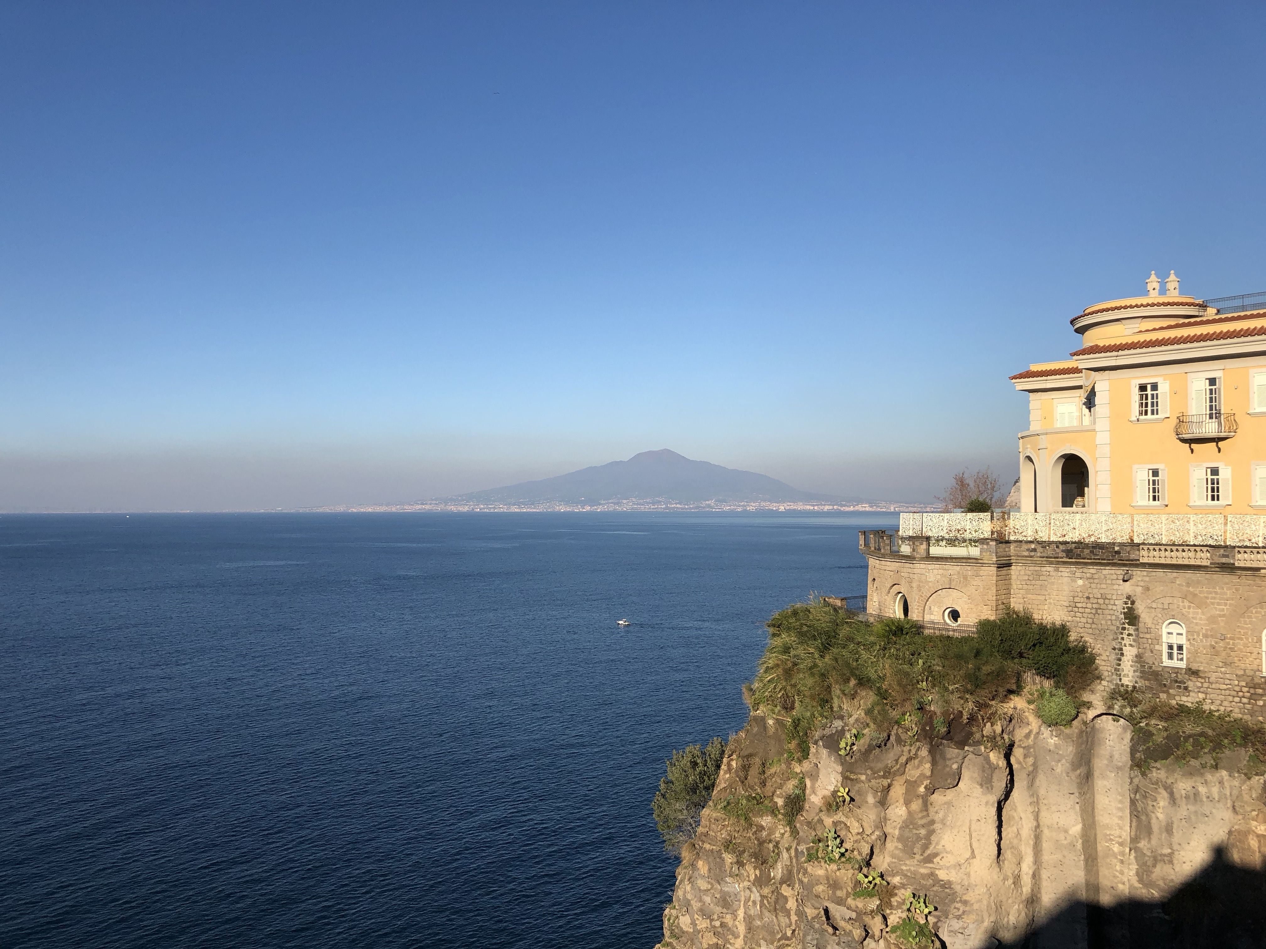 Looking across the bay at Mount Vesuvius, the base of which is  wreathed with brown smog. On the left, perched on a cliff,  is a pale yellow hotel.