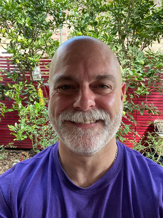 A picture of Nathan. He's a fiftysomething white guy with a shaved head and a white beard. He's standing in a garden in front of a red wooden fence.