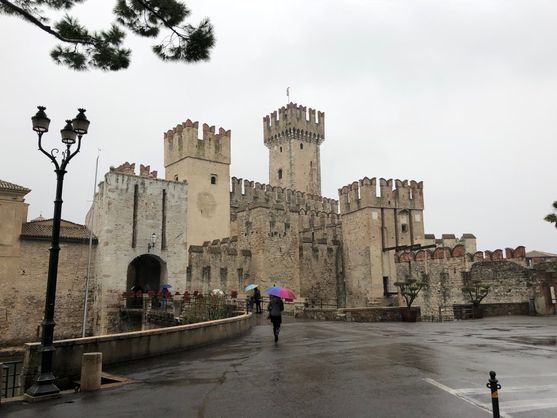 A castle with many square towers with decorative crenellations. It sits atop a bridge over a canal. The road leading up to it is wet with rain, and a couple of tourists are wandering around with brightly coloured umbrellas.