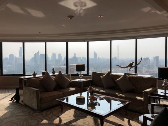 An expensive hotel living room, with a set of couches clustered around a coffee table, lamps, and behind
them a series of windows with a panoramic view of Bangkok