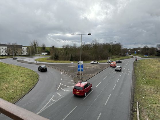 A big roundabout under an overcast sky. There are some halfhearted trees growing in the middle of the roundabout and a few nondescript cars are driving unenthusiastically through it.