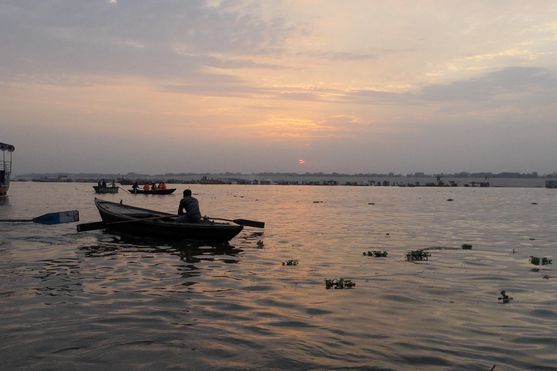 The sun is rising dimly in the distance, lighting up a cloudy sky. On the water we can see a couple of small boats, their hulls and occupants silhouetted against the water.