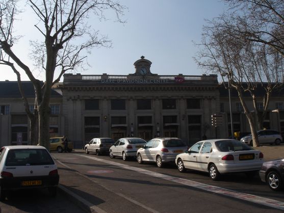 A low nineteenth-century building with the words Gare d'Avignon Centre across the front. There's a car park in front of it.