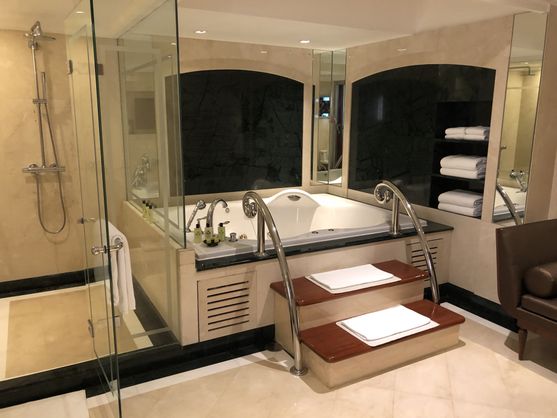 A hotel bathroom, clearly decorated in the 1990s. The centerpiece is a big raised bath with steps leading up to it.