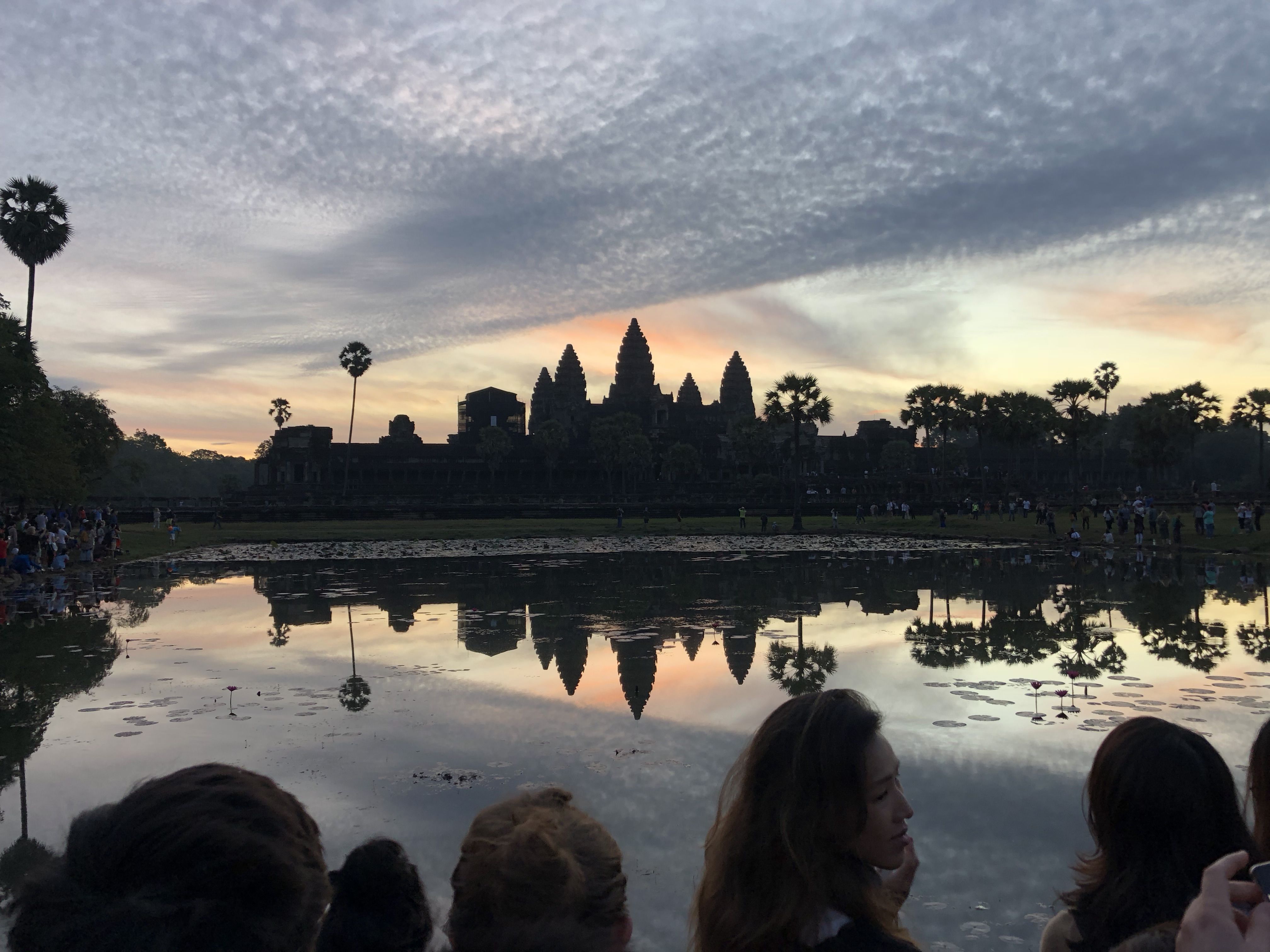 The massive stone temple of Angkor Wat silhouetted against the sunrise and reflected in a vast pool of water in front of it. At the bottom of the photo are the heads of tourists looking at the sight.