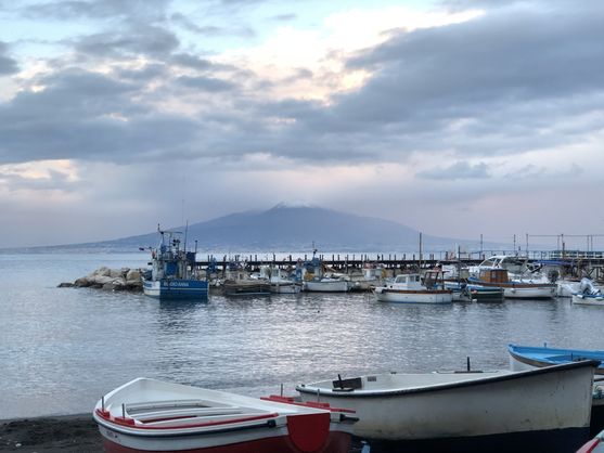 An overcast day at the waterfront at Sorrento. In the foreground are some dinghies pulled up on shore. Behind them is a jetty with boats tied to it. And in the distance is Mount Vesuvius.