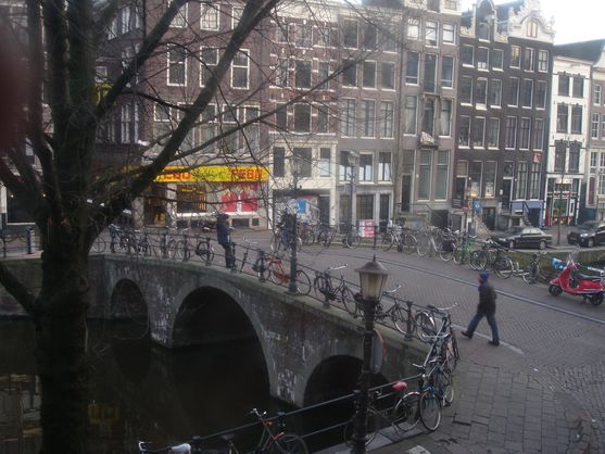 Looking out of the window at a bridge across a canal. There are many bikes leaning against the railings of the bridge. It's cold: people are walking around in coats and hats and the trees have lost their leaves. Lining the canal are tall and narrow Dutch houses.