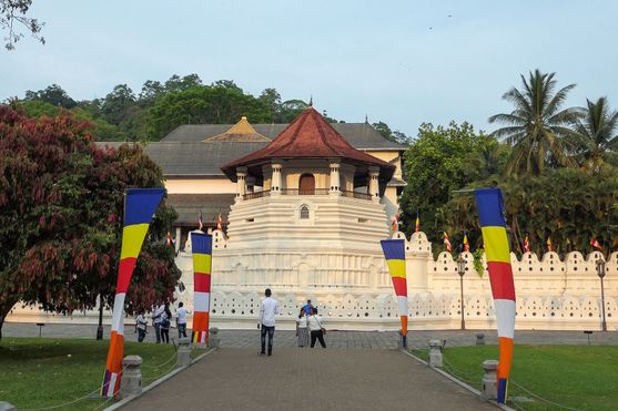 A road lined with Buddhist flags leads to the octagonal turrent of a temple with covered walkway around it and a pointed roof.  In the distance, people have almost reach the temple.