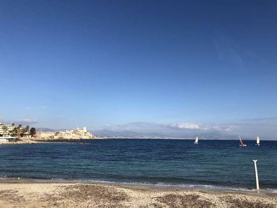 The sky is blue and there are clouds and mountains in the distance as we look across the bay to some white buildings and palm trees. There are a few small yachts on the water.