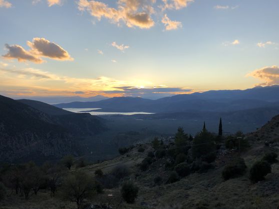 Looking out over a valley at wooded mountains and olive groves, and an inlet in the distance reflecting the light of sunset. Blue and yellow clouds on the horizon.