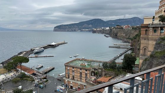 A view of the marina from the top of a cliff in Sorrento. There's a long jetty jutting into the water with a ferry standing nearby. In the distance is a forested headland, closer are some large hotels perched on the top of the cliff.